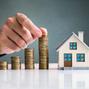 Hand putting a coin on a stack of coins between a scale model house and three smaller stacks of coins
